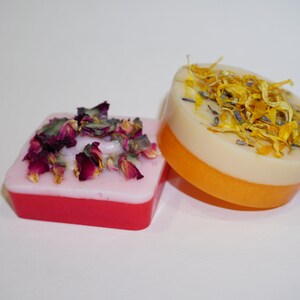 May include: Three handmade soap bars with dried flower petals on top. The soap bars are layered in pink, white, and orange. The soap bars are on a white surface.