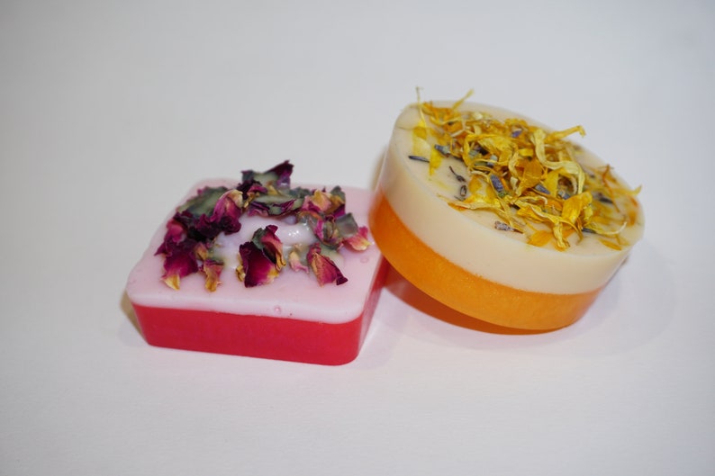 May include: Three handmade soap bars with dried flower petals on top. The soap bars are layered in pink, white, and orange. The soap bars are on a white surface.