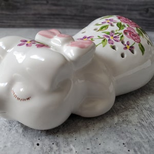 May include: White ceramic pig figurine with pink bow and purple flowers painted on the body. The pig is lying down with its eyes closed.