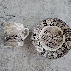 May include: A vintage ceramic mug and plate set. The mug and plate feature a black and white transferware design depicting a waterside scene with buildings and boats. The plate has a floral border.