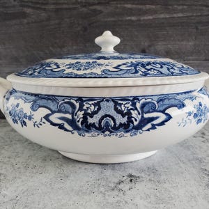 May include: A white and blue ceramic serving dish with a lid. The dish features a blue floral and scroll design. The lid has a white finial. The dish has two handles and is on a gray surface.
