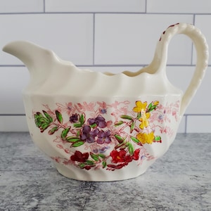 May include: A white ceramic creamer with a floral design in shades of pink, purple, yellow, and green. The creamer has a scalloped edge and a handle.