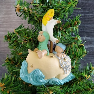 Puede incluir: Un adorno navideño caprichoso con tres pequeñas figuras sobre una ballena color crema con olas azules. Una figura viste un atuendo verde, otra una gorra azul, y la tercera es una luna creciente amarilla con un atuendo blanco.