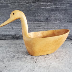 May include: A handcrafted wooden duck-shaped bowl. The bowl is light brown with a long neck and beak, and a hollowed-out body. The duck is carved from a single piece of wood and sits on a gray surface with a dark wood background.