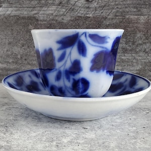 May include: A white teacup and saucer set with a blue floral pattern. The teacup sits on the saucer, both made of ceramic. The blue design features leaves and flowers. The background is a weathered wooden surface.