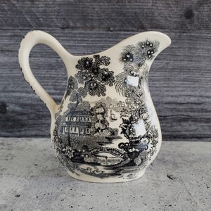 May include: Cream-colored ceramic pitcher with a black and white landscape design. The pitcher features a curved handle and a spout. The landscape design includes trees, buildings, and a bridge. The pitcher is likely for serving beverages.