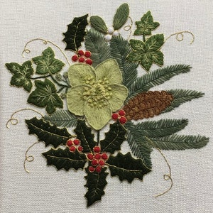 May include: Embroidered floral design with a central pale green flower, dark green holly leaves with red berries, pine needles, and a brown pine cone. Gold thread outlines the leaves and adds detail. The background is a light beige fabric.