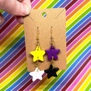 May include: Earrings featuring star-shaped charms in yellow, purple, white, and black. The earrings are suspended from gold-colored hooks and connected with small gold rings. The earrings are displayed on a brown card with a rainbow striped background.