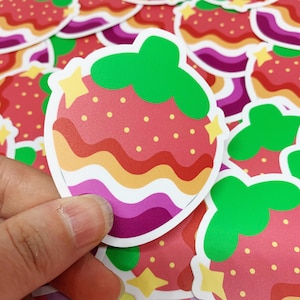 May include: A strawberry sticker with a green stem, red body, and yellow star accents. The strawberry has a wavy design in shades of red, orange, yellow, and purple. The sticker is held in a person's hand, with other similar stickers in the background.