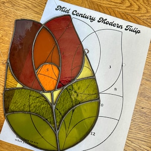 Op de afbeelding: Een gebrandschilderde tulp met rode, oranje en groene glasstukken. De tulp ligt op wit papier met een patroon voor het maken van een gebrandschilderde tulp.