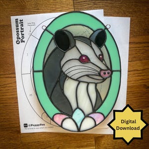 Può includere: Un pannello ovale in vetro colorato raffigurante il ritratto di un opossum con pezzi di vetro nero, bianco e rosa, incorniciato da un bordo verde menta. L'oggetto è appoggiato su un cartamodello con la scritta Opossum Portrait e un badge digitale.