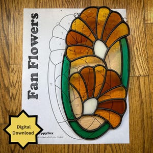 May include: A stained glass panel featuring two fan-shaped flowers in shades of amber and orange with white centers and green borders, displayed over a printed paper pattern labeled Fan Flowers with a yellow star icon stating Digital Download.