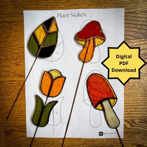 May include: Four stained glass plant stakes with a digital download label. The stakes are shaped like a leaf, a mushroom, a tulip, and a mushroom. The leaf is green, yellow, and black. The mushrooms are red and orange. The tulip is orange and yellow. The stakes are on a white background.