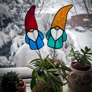 Two garden gnome wizards stained glass plant stakes displayed in a hoya plant on a windowsill. Outside the window is a bush with a lot of fresh white snow. The gnome stained glass garden stakes are wearing long, tall hats white beards and blue coats. Mid Century Modern (MCM) / Bohemian / Retro / Vintage style from the 1970s or 1960s. The colors are red, blue, white, teal and orange