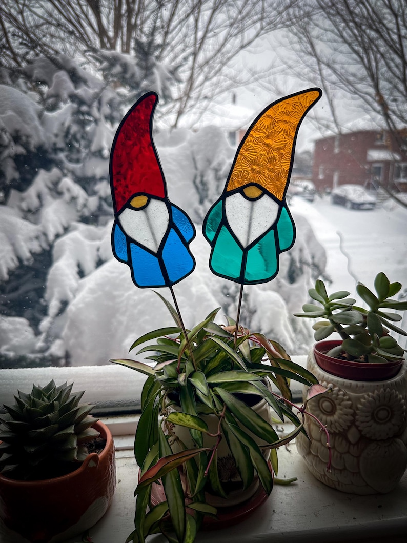 Two garden gnome wizards stained glass plant stakes displayed in a hoya plant on a windowsill. Outside the window is a bush with a lot of fresh white snow. The gnome stained glass garden stakes are wearing long, tall hats white beards and blue coats. Mid Century Modern (MCM) / Bohemian / Retro / Vintage style from the 1970s or 1960s. The colors are red, blue, white, teal and orange