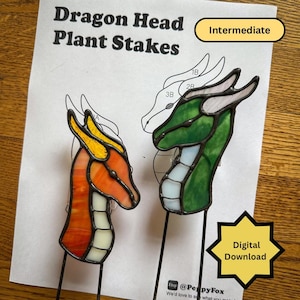 May include: Two stained glass dragon head plant stakes. One is orange and yellow, the other is green and white. The text "Dragon Head Plant Stakes" is at the top of the image, with "Intermediate" and "Digital Download" labels.