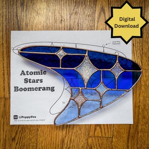 May include: A stained glass boomerang design with the text "Atomic Stars Boomerang." The design features shades of blue and clear glass, outlined with copper. A digital download icon is in the upper right corner.