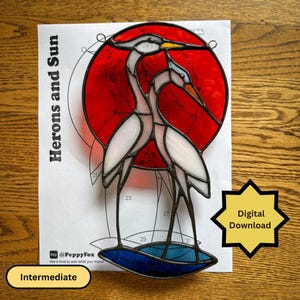 Op de afbeelding: Een glas-in-lood suncatcher patroon getiteld Herons and Sun, met twee witte reigers tegen een grote rode cirkelvormige zon en een blauwe basis. Het ontwerp bevat genummerde secties voor montage, tekstlabels en een gele Digital Download-ster.