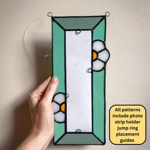 May include: A rectangular stained glass photo strip holder featuring a mint green border and two white flower motifs with orange centers. The frame includes metal jump rings for hanging and a central white paper insert with the text DNP repeated.