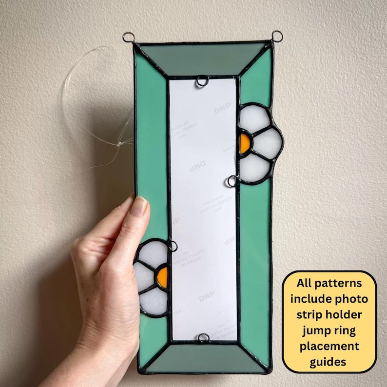 May include: A rectangular stained glass photo strip holder featuring a mint green border and two white flower motifs with orange centers. The frame includes metal jump rings for hanging and a central white paper insert with the text DNP repeated.