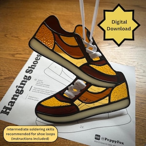 Op de afbeelding: Hangende schoenen van glas in lood in bruin-, geel- en goudtinten, met witte veters. De schoenen worden opgehangen met witte koorden. De afbeelding bevat een digitale downloadafbeelding en instructies voor schoenlusjes.