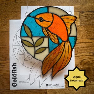 May include: A circular stained glass goldfish suncatcher featuring orange, blue, and olive green glass pieces held by black lead lines. The piece is displayed over a printed paper pattern labeled Goldfish with Digital Download text.