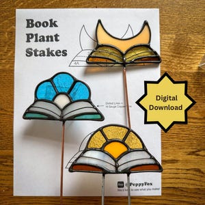 May include: Three stained glass book plant stakes with copper stems. One features a fox design, another a blue flower, and the third a yellow sun. The stakes are on a wooden surface with a paper that says "Book Plant Stakes" and "Digital Download".
