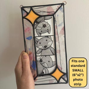 May include: A rectangular stained glass frame with black metal soldering, featuring two orange star-shaped accents at the corners and a clear glass border with pink and blue confetti-like inclusions. It holds a vertical strip of three cartoon foxes.