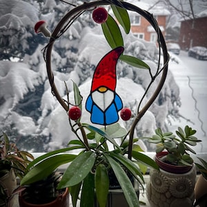 A garden gnome wizards stained glass plant stakes displayed in a hoya plant on a windowsill. Outside the window is a bush with a lot of fresh white snow. The gnome stained glass garden stake is wearing a long, tall, red hat; has a white white beards and a blue coat. Mid Century Modern (MCM) / Bohemian / Retro / Vintage style from the 1970s or 1960s. The colors are red, blue, white, teal and orange