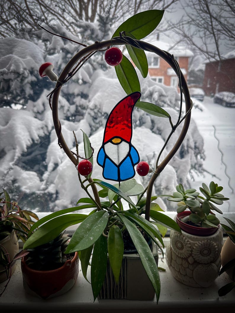 A garden gnome wizards stained glass plant stakes displayed in a hoya plant on a windowsill. Outside the window is a bush with a lot of fresh white snow. The gnome stained glass garden stake is wearing a long, tall, red hat; has a white white beards and a blue coat. Mid Century Modern (MCM) / Bohemian / Retro / Vintage style from the 1970s or 1960s. The colors are red, blue, white, teal and orange