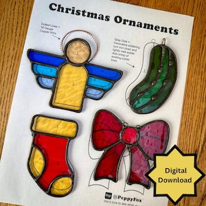 Puede incluir: Una hoja con instrucciones para hacer adornos navideños de vidrieras. Los diseños incluyen un ángel, un pepinillo, un calcetín y un lazo. El ángel tiene alas azules y amarillas. El lazo es rojo y rosa. El texto "Christmas Ornaments" está en la parte superior.