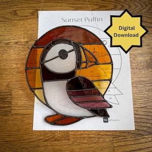 May include: Stained glass artwork of a puffin bird with sunset colors. The bird has a white chest, a black eye, and a colorful head with shades of orange, yellow, and brown. The words "Sunset Puffin" are above the bird.