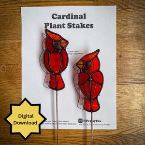 May include: Two red cardinal plant stakes with copper wire stems are displayed on a wooden surface. The cardinals are made of stained glass with black outlines. The text "Cardinal Plant Stakes" is printed above the cardinals.