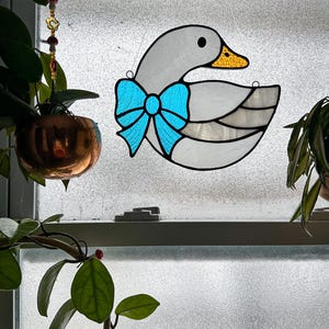 May include: A stained glass goose with a light blue bow. The goose has a grey body, a yellow beak, and a black eye. The stained glass is hanging in front of a window.