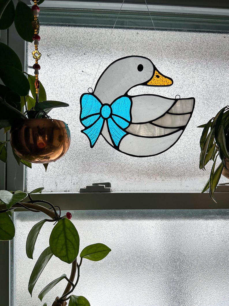 May include: A stained glass goose with a light blue bow. The goose has a grey body, a yellow beak, and a black eye. The stained glass is hanging in front of a window.