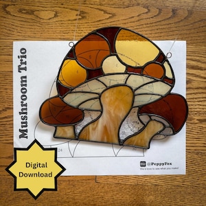 May include: A stained glass mushroom trio in shades of brown, yellow, and cream. The design is on a white paper with the text "Mushroom Trio" and "Digital Download". The image is on a wooden surface.