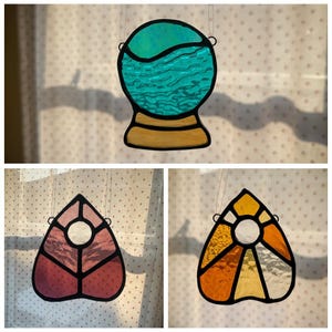 May include: Three stained glass suncatchers. The top one is a crystal ball with blue and turquoise water. The bottom left is a pink leaf shape. The bottom right is a yellow and orange sunburst design. All have black outlines.