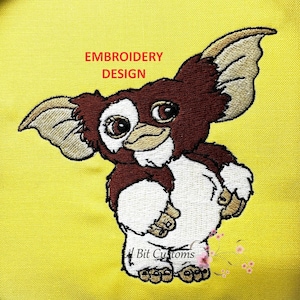 May include: Embroidered design of a brown and white furry creature with large ears, big eyes, and a small beak. The design is on a yellow background and includes the text "Embroidery Design".
