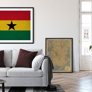 May include: A framed print of the flag of Ghana, featuring a red, yellow, and green horizontal tricolor with a black five-pointed star in the center. The print is hanging on a white wall above a white couch with two burgundy pillows. A framed map is hanging on the wall to the right of the flag print.