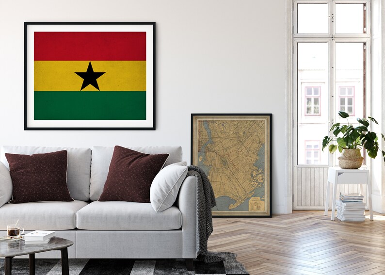 May include: A framed print of the flag of Ghana, featuring a red, yellow, and green horizontal tricolor with a black five-pointed star in the center. The print is hanging on a white wall above a white couch with two burgundy pillows. A framed map is hanging on the wall to the right of the flag print.