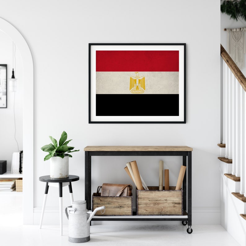 May include: A framed print of the flag of Egypt. The flag is red, white, and black with a gold eagle in the center. The flag is displayed on a white wall above a wooden console table.