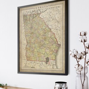 May include: Vintage map print of the state of Georgia, framed in black. The map is in shades of yellow, green, and brown, with red lines indicating roads and railroads. The title "GEORGIA" is printed at the top of the map.