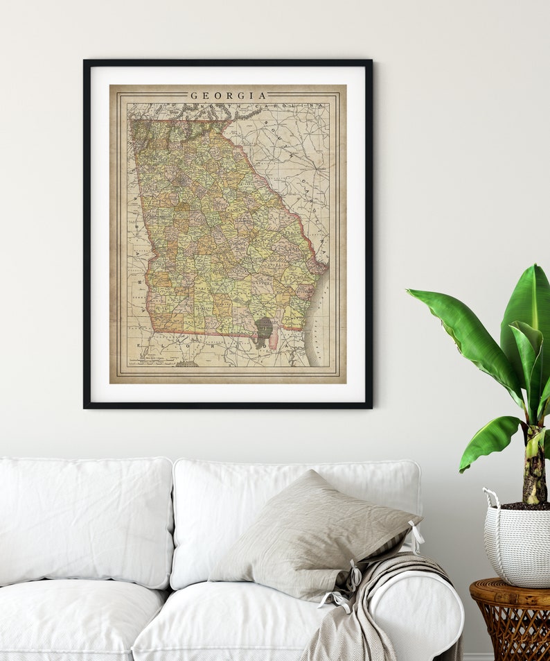 May include: A vintage map of Georgia, printed in black, brown, yellow, and green on a cream-colored background. The map is framed in a black frame and hanging on a white wall.
