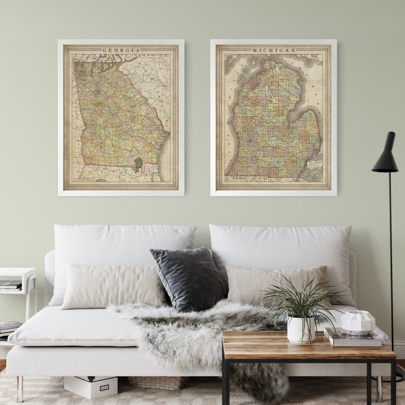 May include: Two vintage maps of Georgia and Michigan in white frames are displayed on a light green wall above a white couch with a faux fur throw and a wooden coffee table.