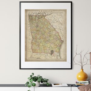 May include: A vintage map of Georgia, printed in black, brown, yellow, and green, with the state name "GEORGIA" printed at the top in black lettering. The map is framed in a black frame and hanging on a white wall.