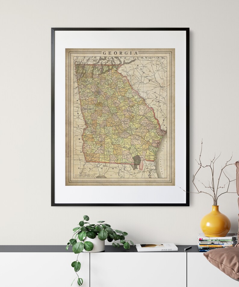 May include: A vintage map of Georgia, printed in black, brown, yellow, and green, with the state name "GEORGIA" printed at the top in black lettering. The map is framed in a black frame and hanging on a white wall.