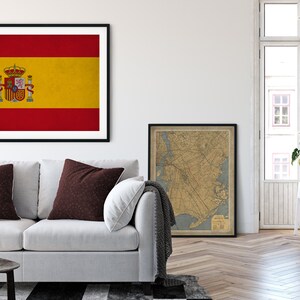 May include: Framed print of the Spanish flag, featuring red and yellow horizontal stripes with the Spanish coat of arms. The artwork hangs above a white sofa with brown accent pillows. A vintage map is also framed.
