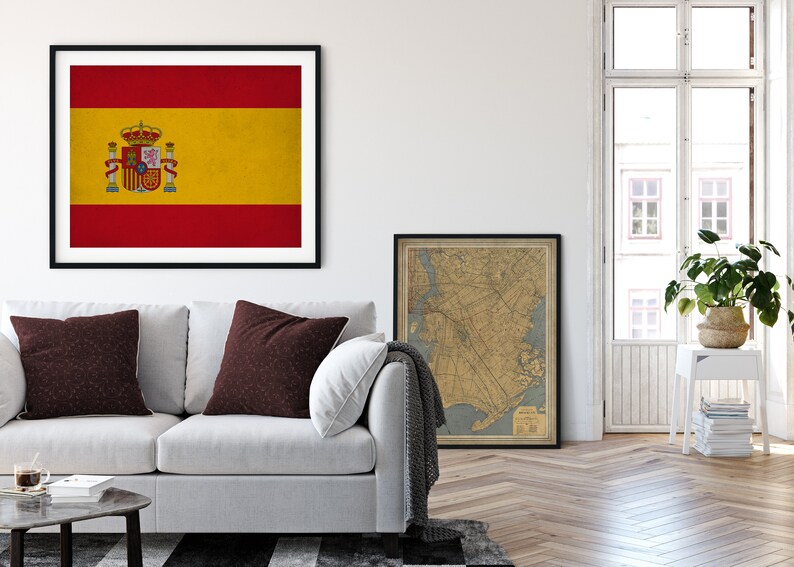 May include: Framed print of the Spanish flag, featuring red and yellow horizontal stripes with the Spanish coat of arms. The artwork hangs above a white sofa with brown accent pillows. A vintage map is also framed.
