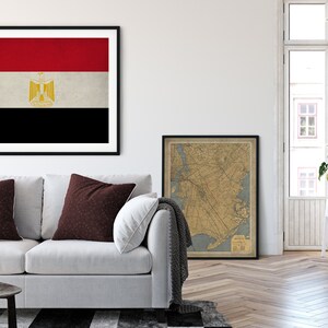 May include: A white couch with two maroon pillows in a living room setting. A framed print of the flag of Egypt hangs on the wall above the couch. A framed vintage map hangs on the wall to the right of the couch. A white side table with a potted plant and a stack of books sits in the corner of the room.