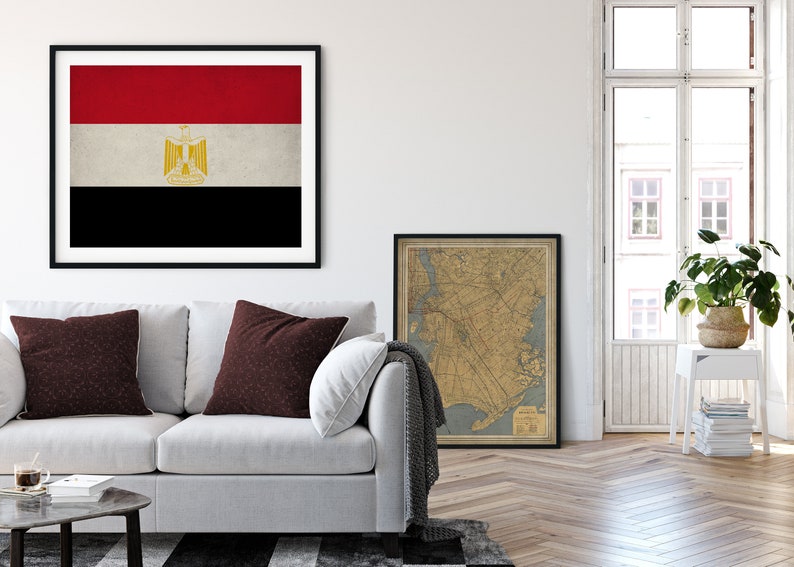 May include: A white couch with two maroon pillows in a living room setting. A framed print of the flag of Egypt hangs on the wall above the couch. A framed vintage map hangs on the wall to the right of the couch. A white side table with a potted plant and a stack of books sits in the corner of the room.